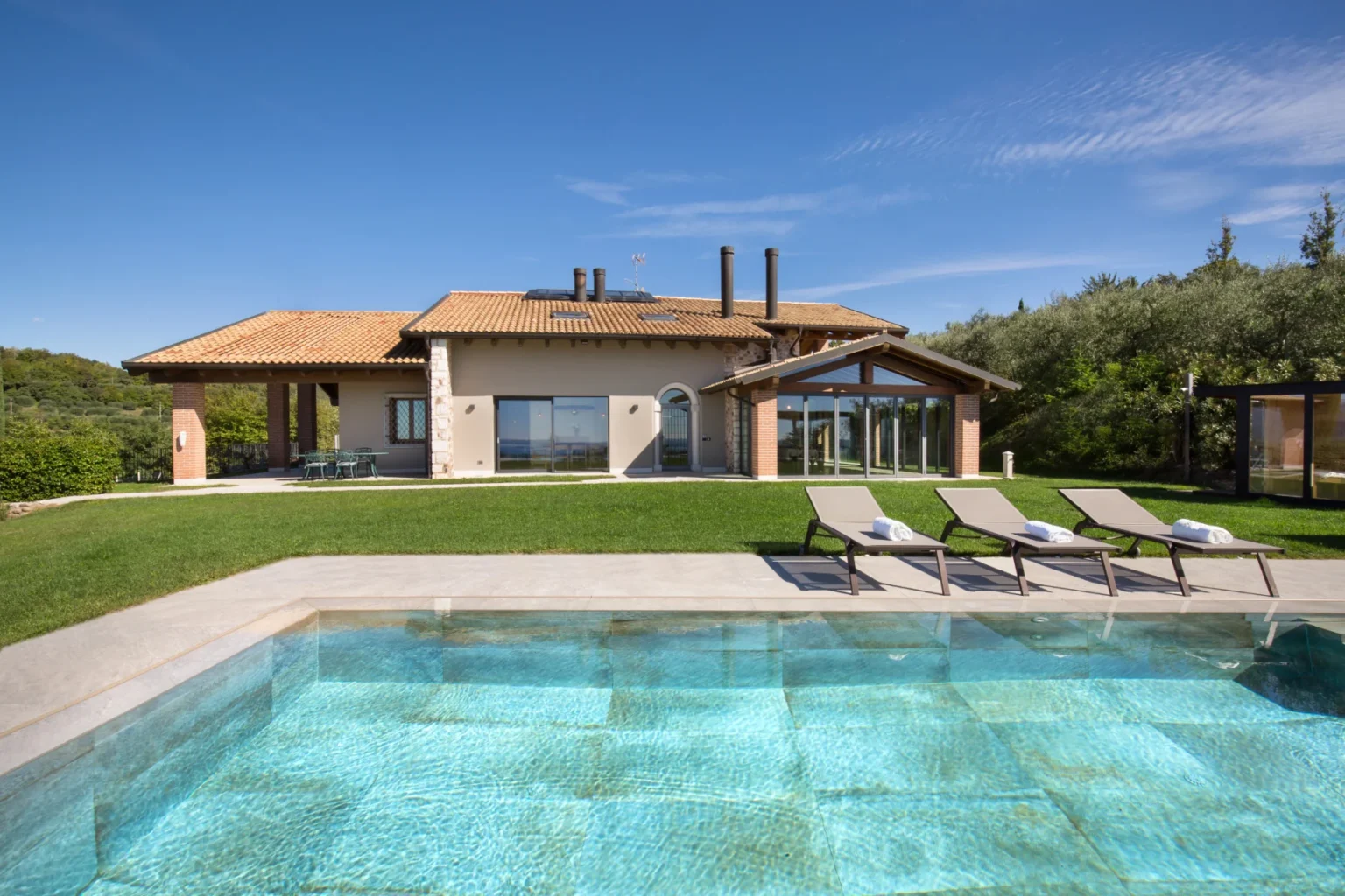 Modern villa with a swimming pool, surrounded by lush greenery, featuring a spacious lawn and outdoor seating area. The house showcases a combination of stone and contemporary architecture under a clear blue sky. | Only on SopranoVillas