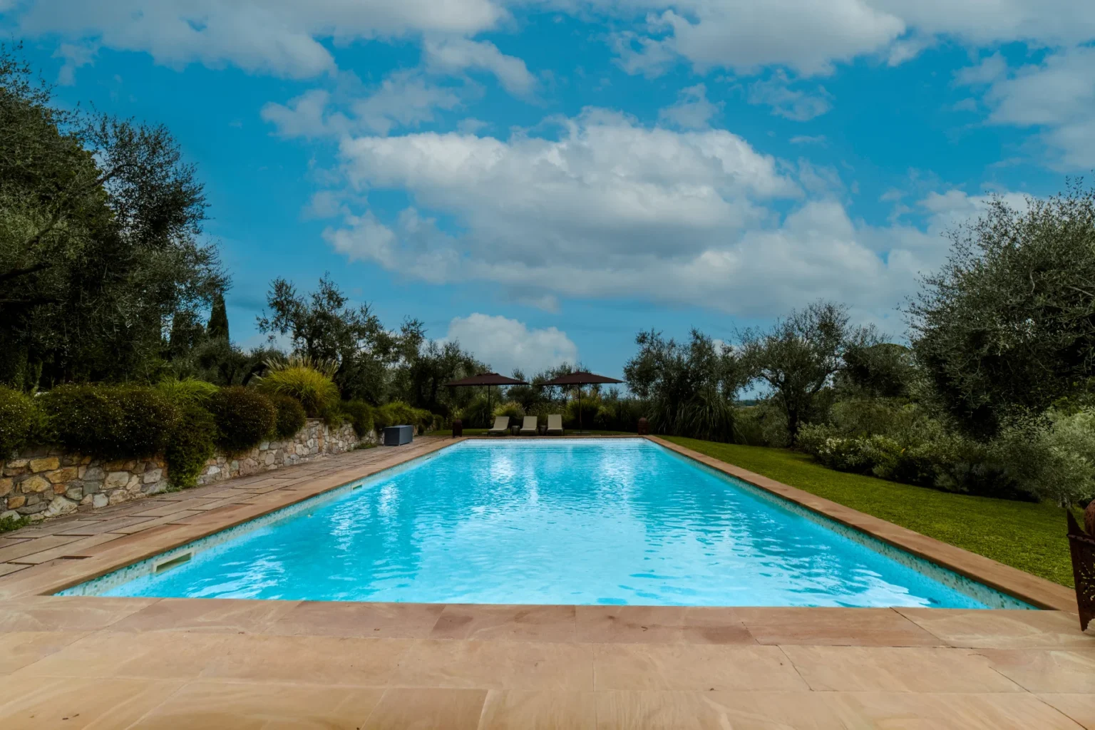Serene outdoor swimming pool surrounded by lush greenery and olive trees under a blue sky with fluffy clouds, featuring comfortable lounge chairs and a stone pathway. | Only on SopranoVillas