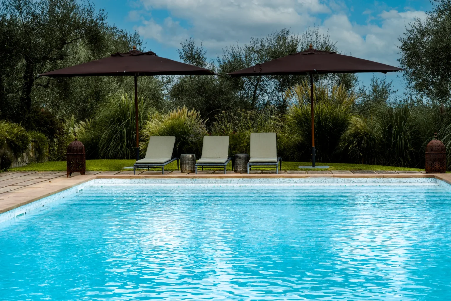 Inviting outdoor pool area featuring three lounge chairs under large umbrellas, surrounded by lush greenery and a serene landscape, perfect for relaxation and leisure. | Only on SopranoVillas