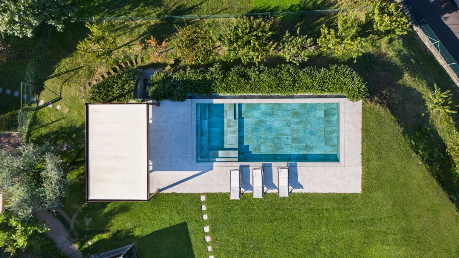 Aerial view of a modern swimming pool surrounded by lush greenery, featuring a spacious deck with lounge chairs and a shaded area, set in a well-maintained garden. | Only on SopranoVillas