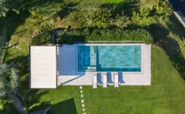 Aerial view of a modern swimming pool surrounded by lush greenery, featuring a spacious deck with lounge chairs and a shaded area, set in a well-maintained garden.