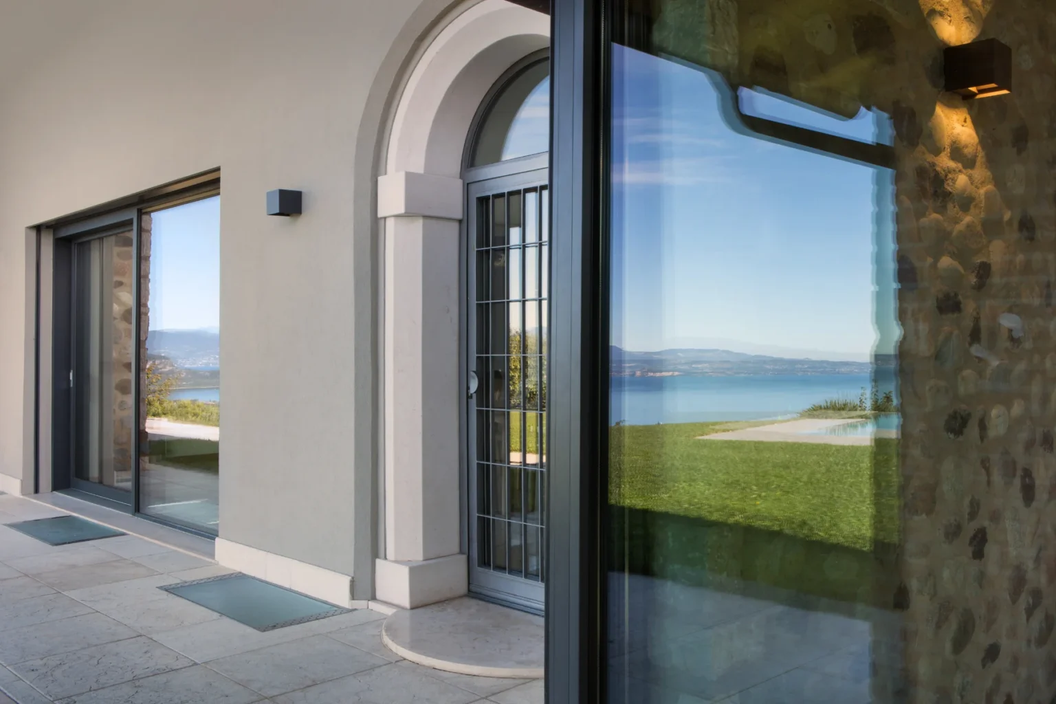 Modern entrance of a luxury home featuring large glass doors, a stone wall, and a scenic view of a lake and mountains in the background. The architecture showcases a blend of contemporary design and natural elements. | Only on SopranoVillas