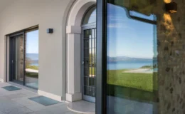Modern entrance of a luxury home featuring large glass doors, a stone wall, and a scenic view of a lake and mountains in the background. The architecture showcases a blend of contemporary design and natural elements.