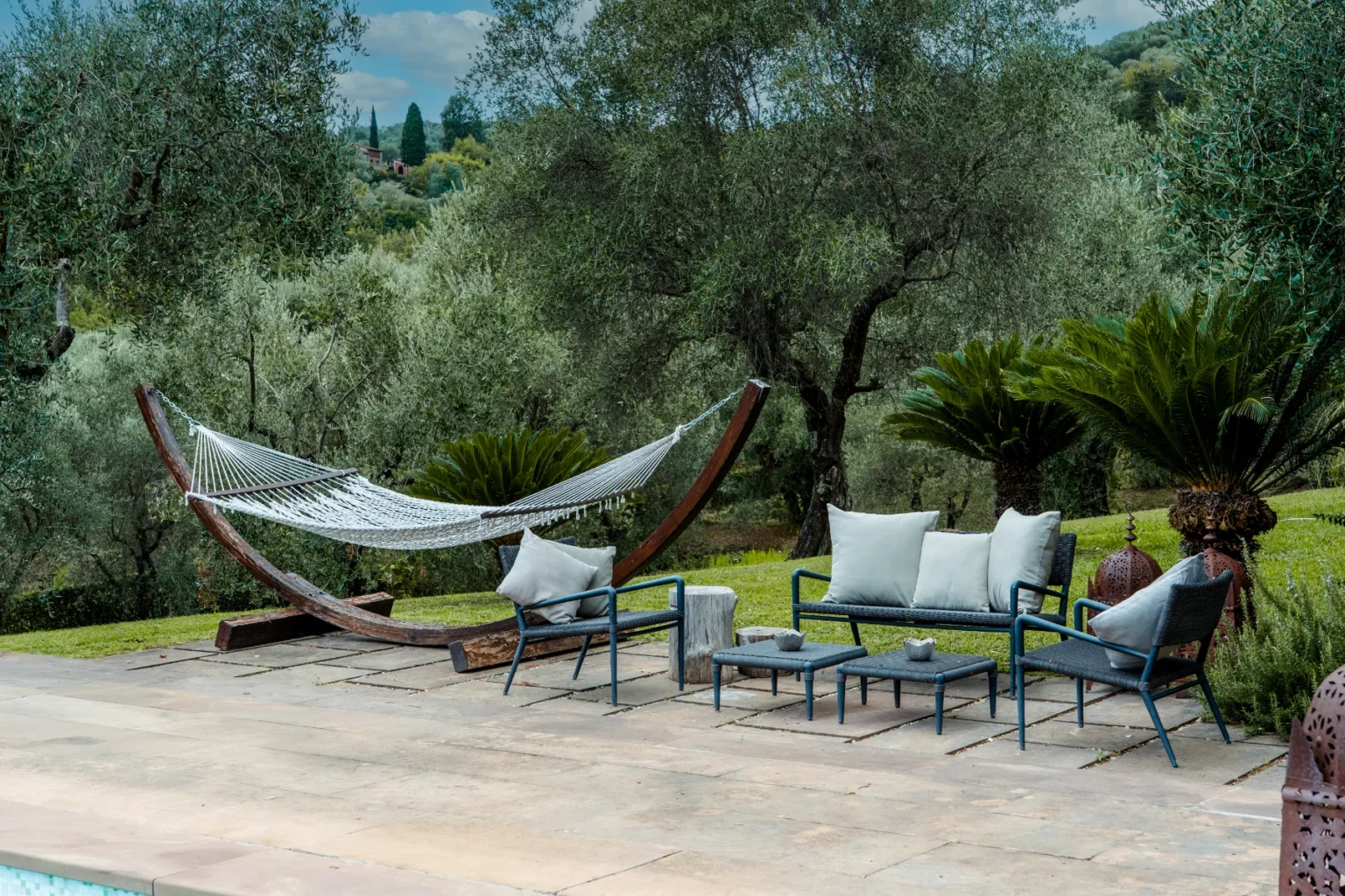 Cozy outdoor seating area featuring a hammock, modern chairs, and a coffee table surrounded by lush olive trees and greenery, perfect for relaxation and enjoying nature. | Only on SopranoVillas
