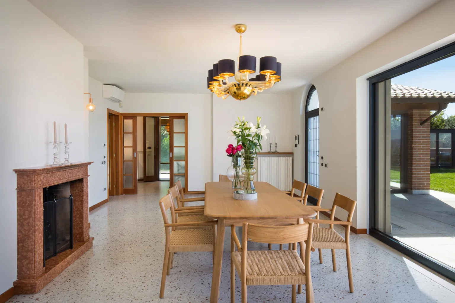 Modern dining room with a wooden table and six chairs, featuring a stylish chandelier and a decorative vase with flowers. The room has large windows providing natural light and a view of the garden, with a marble fireplace adding elegance to the space. | Only on SopranoVillas