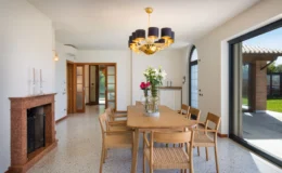 Modern dining room with a wooden table and six chairs, featuring a stylish chandelier and a decorative vase with flowers. The room has large windows providing natural light and a view of the garden, with a marble fireplace adding elegance to the space.