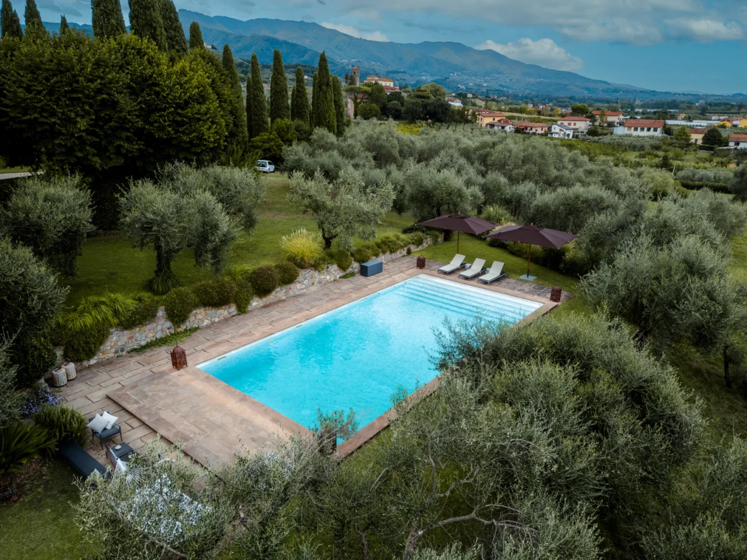 Aerial view of a serene swimming pool surrounded by lush olive trees and manicured gardens, with lounge chairs and umbrellas for relaxation, set against a backdrop of rolling hills and a scenic landscape. | Only on SopranoVillas