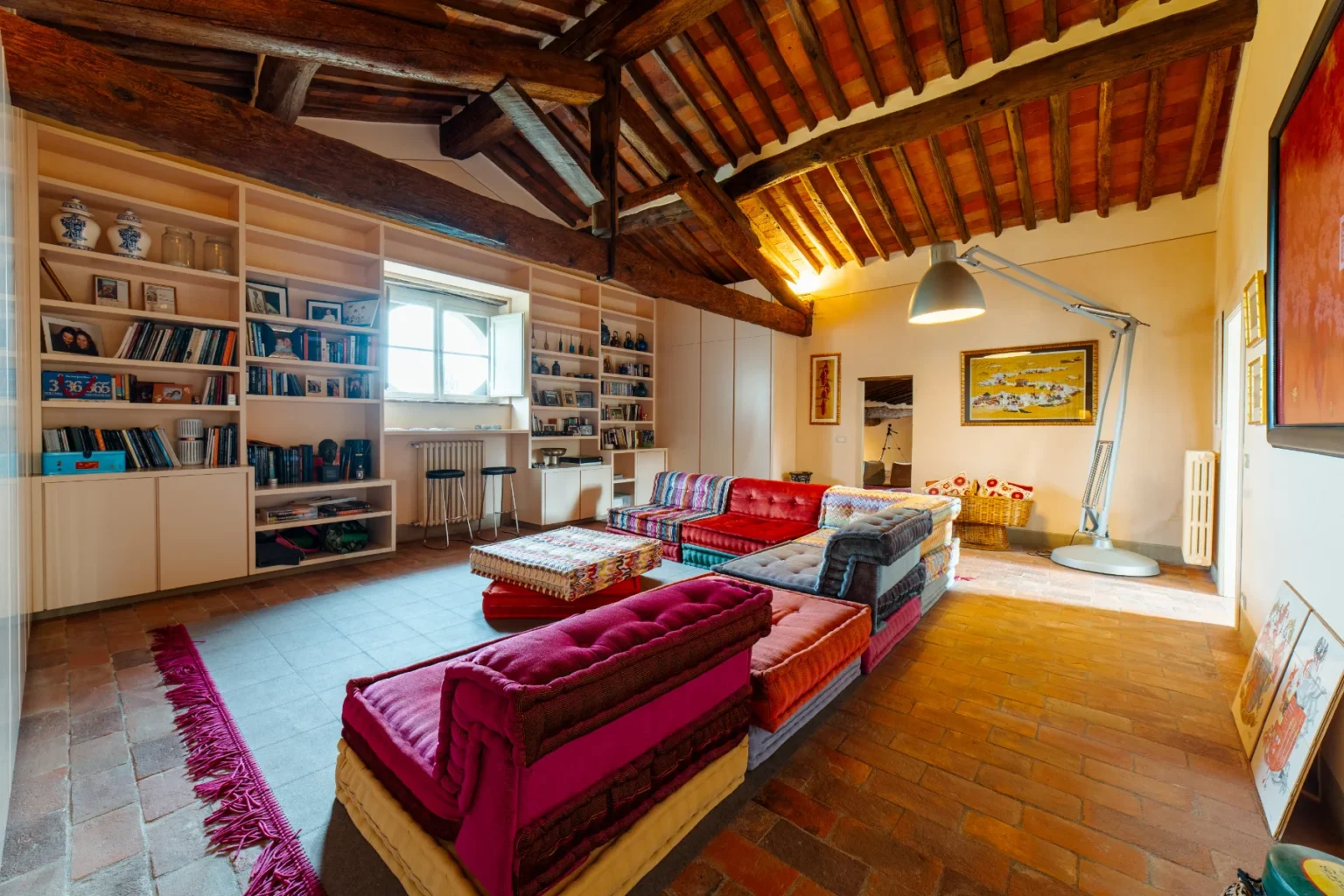 Spacious living room featuring high wooden ceilings, colorful seating arrangements, and a large bookshelf filled with books and decorative items. Natural light floods the space through a window, highlighting the warm tones of the brick floor and cozy decor. Ideal for relaxation and gatherings. | Only on SopranoVillas
