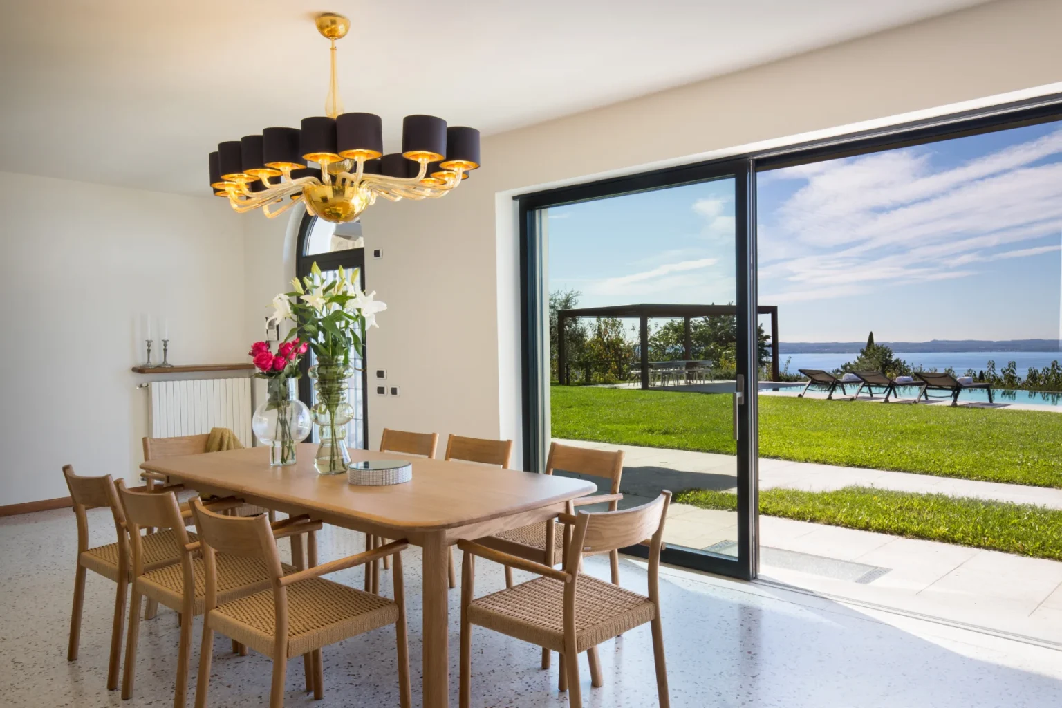Modern dining room featuring a wooden table with woven chairs, a decorative chandelier, and large glass doors opening to a scenic outdoor view with a pool and lawn. Fresh flowers in vases add a touch of elegance to the space. | Only on SopranoVillas