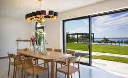 Modern dining room featuring a wooden table with woven chairs, a decorative chandelier, and large glass doors opening to a scenic outdoor view with a pool and lawn. Fresh flowers in vases add a touch of elegance to the space.