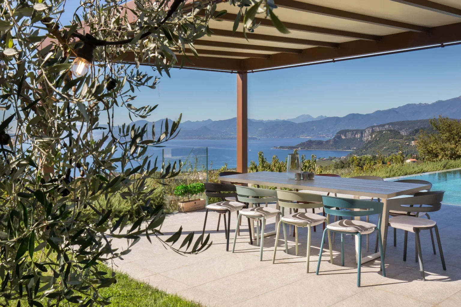Outdoor dining area with a modern table and colorful chairs overlooking a scenic view of mountains and a lake, framed by olive branches and a pergola. | Only on SopranoVillas