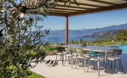 Outdoor dining area with a modern table and colorful chairs overlooking a scenic view of mountains and a lake, framed by olive branches and a pergola.