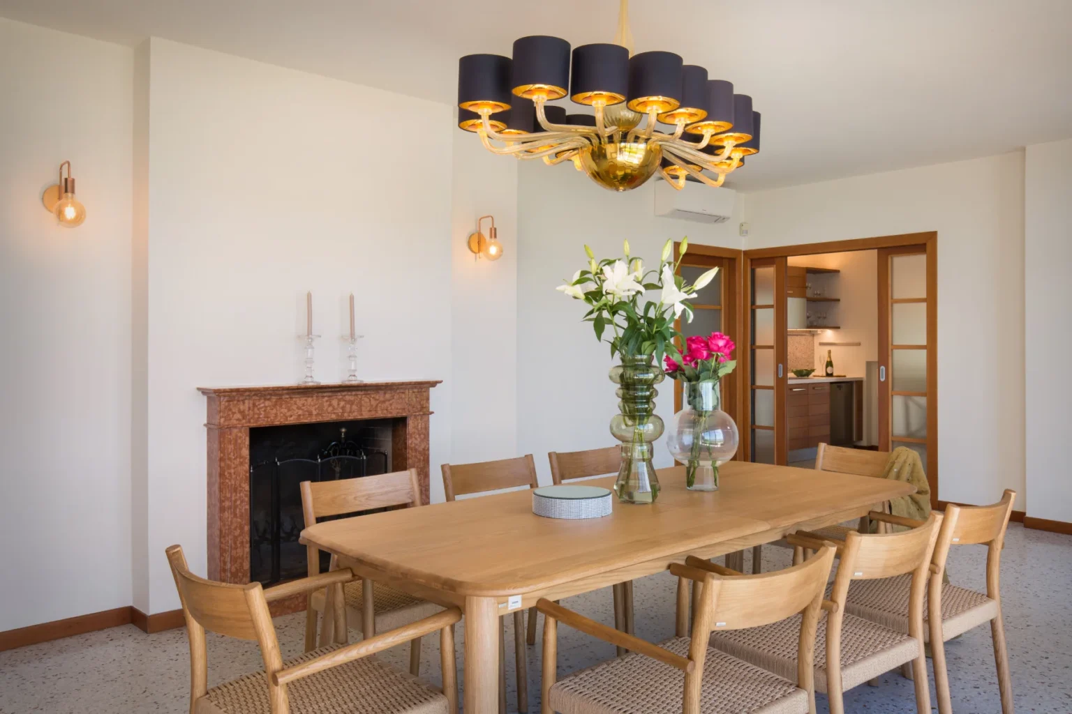 Bright and modern dining room featuring a large wooden table surrounded by stylish chairs, a vibrant floral centerpiece, and an elegant chandelier. A marble fireplace adds warmth to the space, complemented by soft wall lighting and a glimpse of a contemporary kitchen in the background. | Only on SopranoVillas
