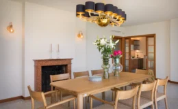 Bright and modern dining room featuring a large wooden table surrounded by stylish chairs, a vibrant floral centerpiece, and an elegant chandelier. A marble fireplace adds warmth to the space, complemented by soft wall lighting and a glimpse of a contemporary kitchen in the background.