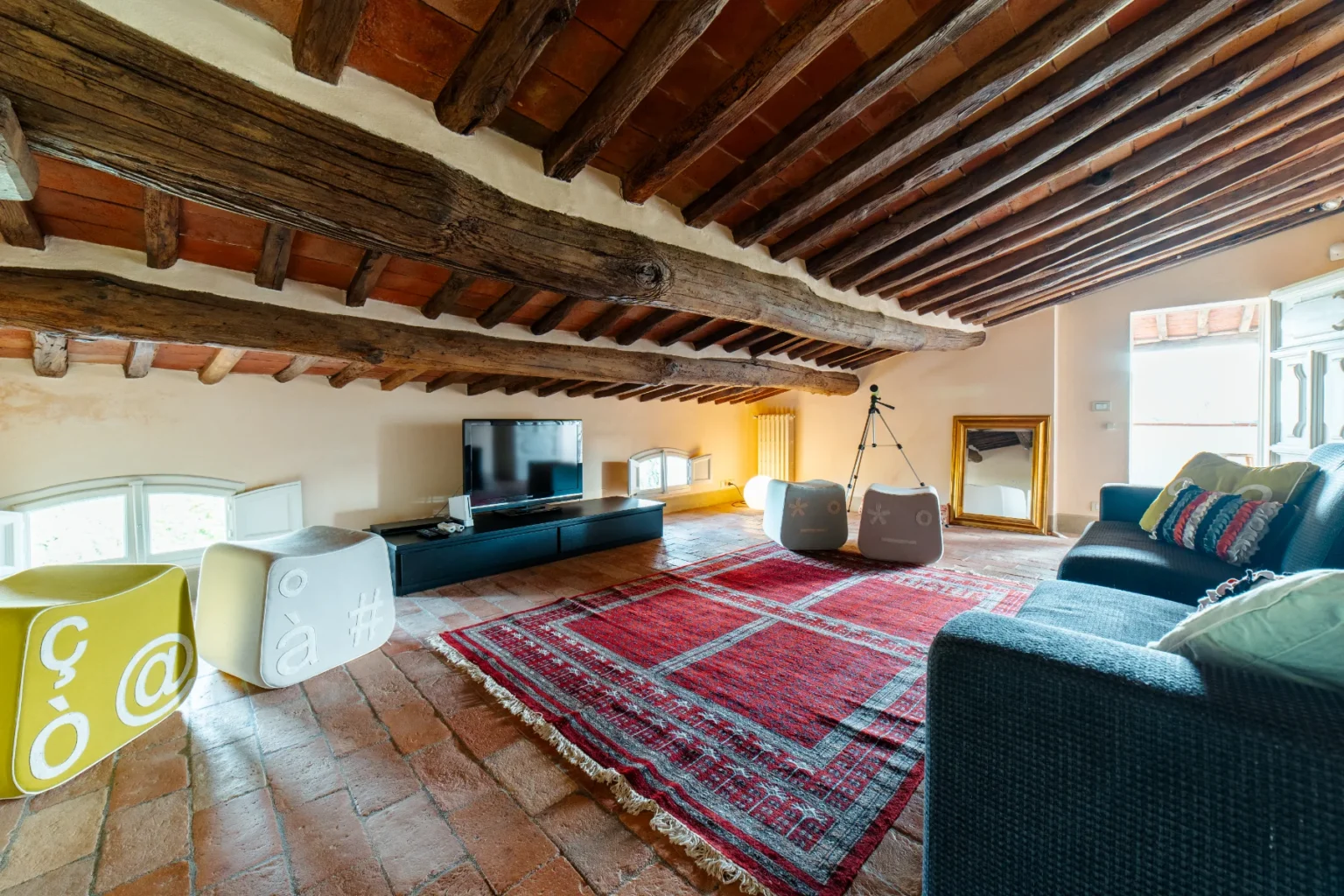 Cozy attic living room featuring wooden beams, terracotta flooring, and a vintage rug, with a television, modern seating, and unique decorative stools in vibrant colors. Natural light fills the space from a window, creating a warm and inviting atmosphere. | Only on SopranoVillas