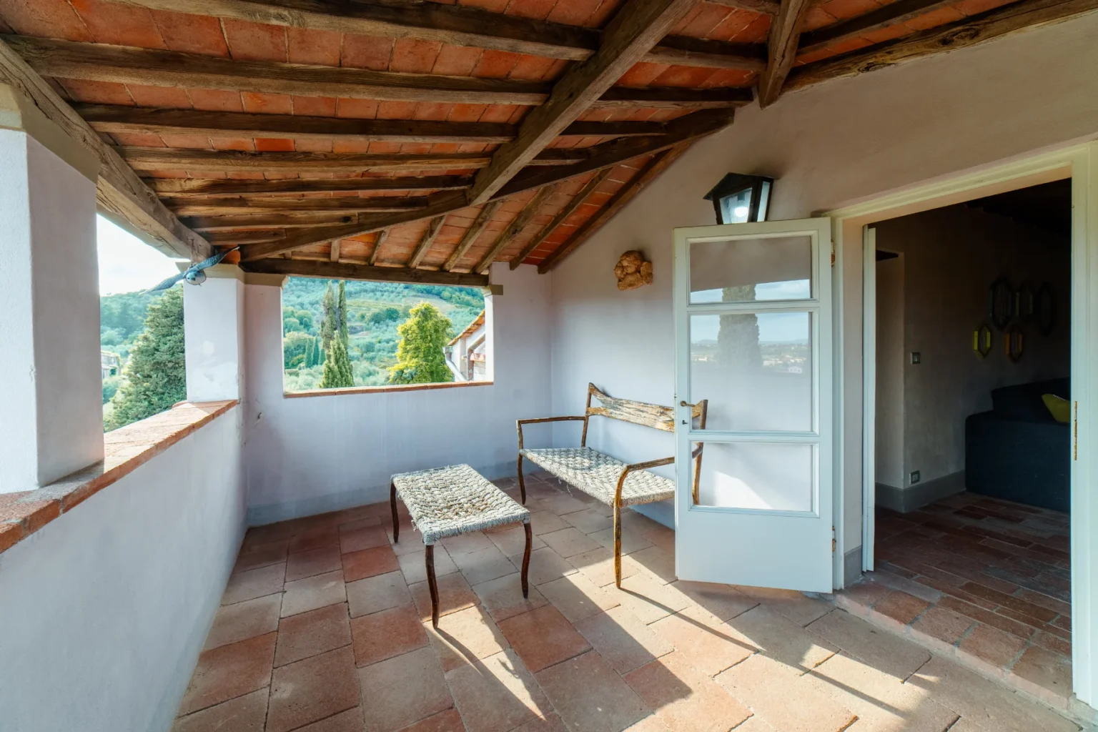 Cozy balcony with rustic wooden beams and terracotta tiles, featuring a woven bench and table, offering a scenic view of lush greenery and hills. Natural light floods the space through open windows, creating a serene outdoor retreat. | Only on SopranoVillas