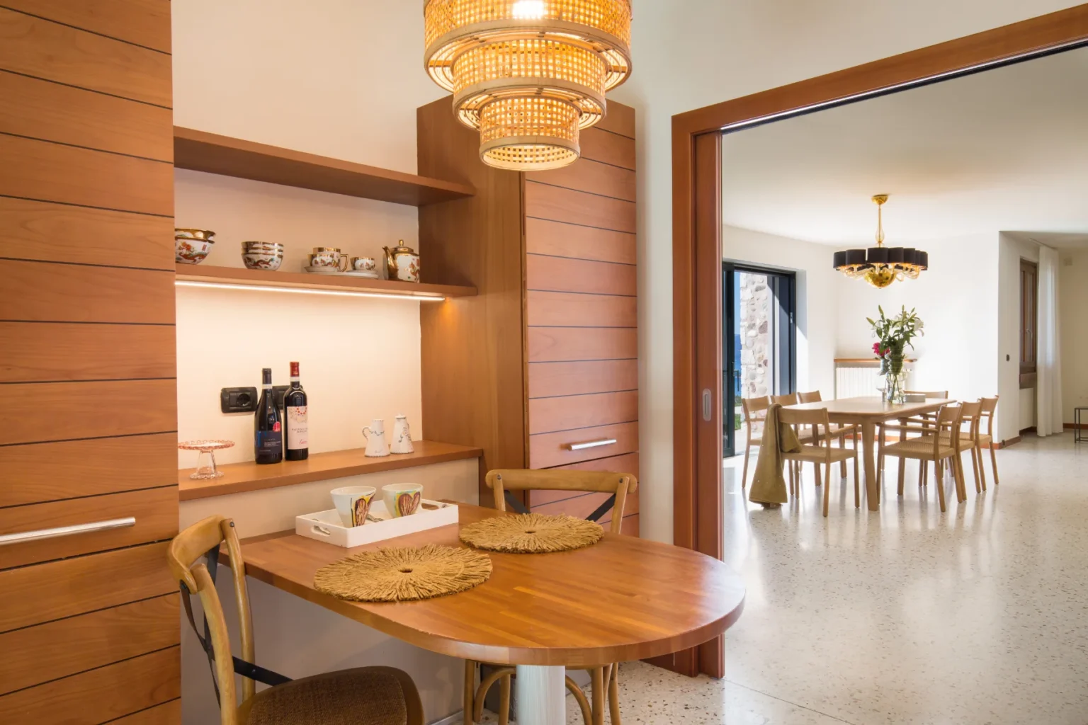 Modern kitchen and dining area featuring a wooden table with decorative placemats, a shelf displaying fine china and wine bottles, and a stylish chandelier. The open layout connects to a spacious dining room with a large table and floral centerpiece. Ideal for entertaining and everyday dining. | Only on SopranoVillas