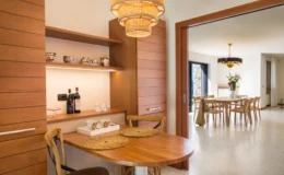 Modern kitchen and dining area featuring a wooden table with decorative placemats, a shelf displaying fine china and wine bottles, and a stylish chandelier. The open layout connects to a spacious dining room with a large table and floral centerpiece. Ideal for entertaining and everyday dining.