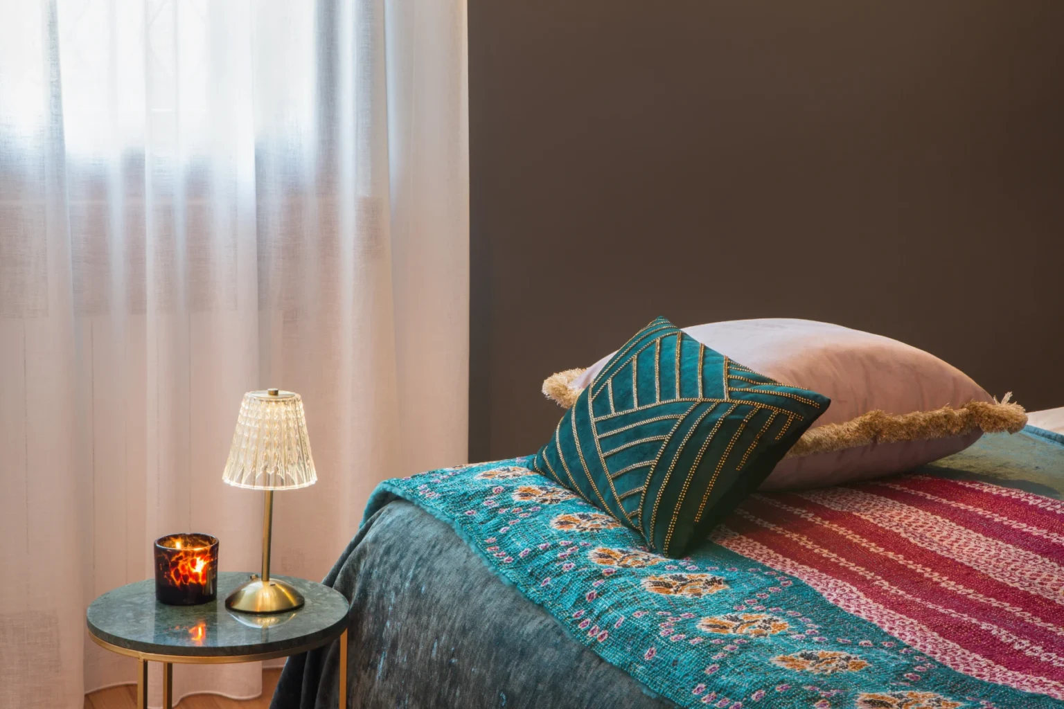 Cozy bedroom corner featuring a colorful quilt, decorative pillows, a small bedside table with a candle, and a stylish lamp, illuminated by natural light from sheer curtains. | Only on SopranoVillas