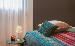 Cozy bedroom corner featuring a colorful quilt, decorative pillows, a small bedside table with a candle, and a stylish lamp, illuminated by natural light from sheer curtains.