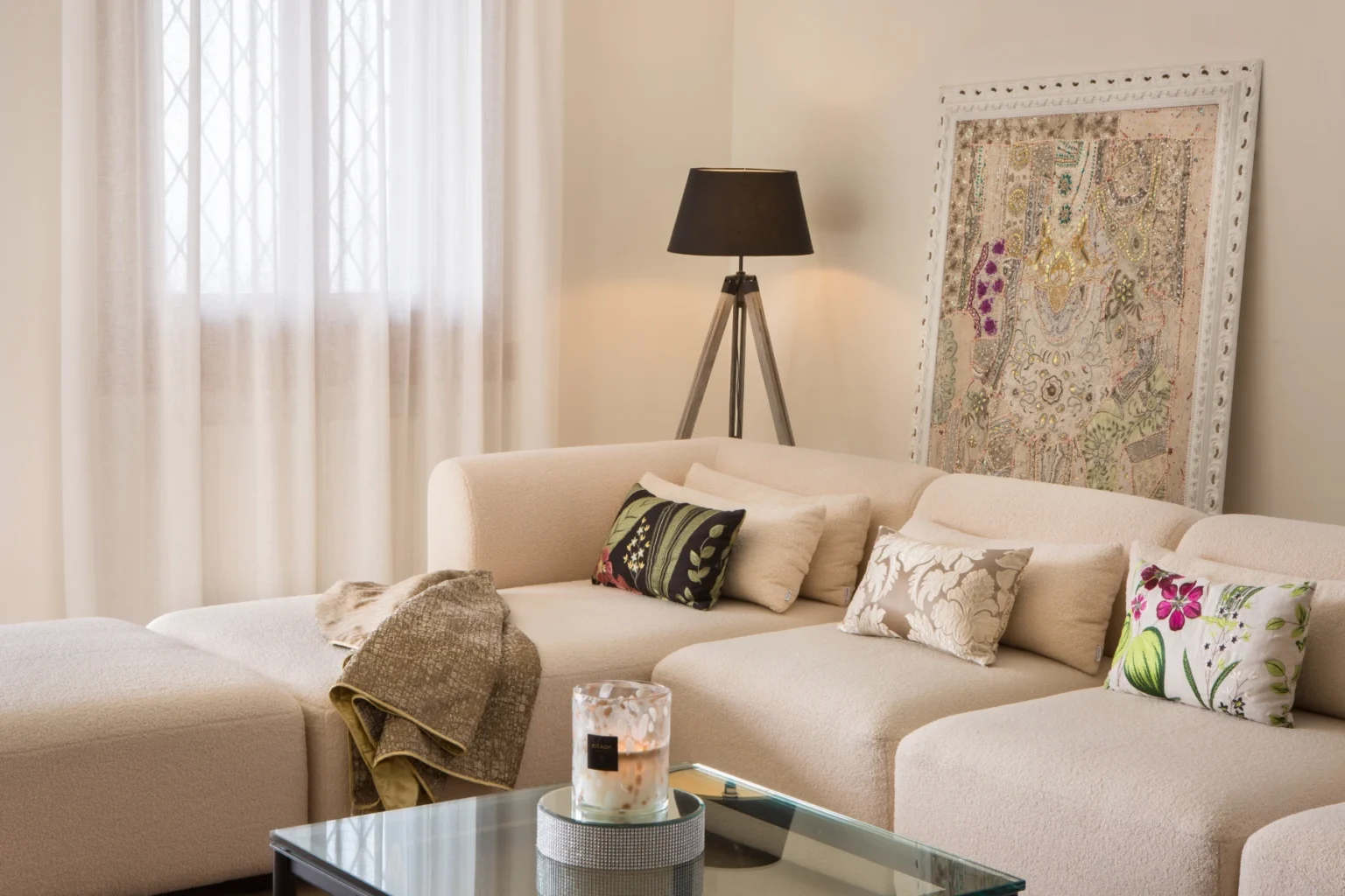 Cozy modern living room featuring a beige sectional sofa with decorative pillows, a stylish floor lamp, a glass coffee table, and a colorful wall art piece, illuminated by natural light from a window. | Only on SopranoVillas