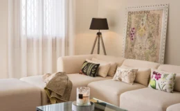 Cozy modern living room featuring a beige sectional sofa with decorative pillows, a stylish floor lamp, a glass coffee table, and a colorful wall art piece, illuminated by natural light from a window.