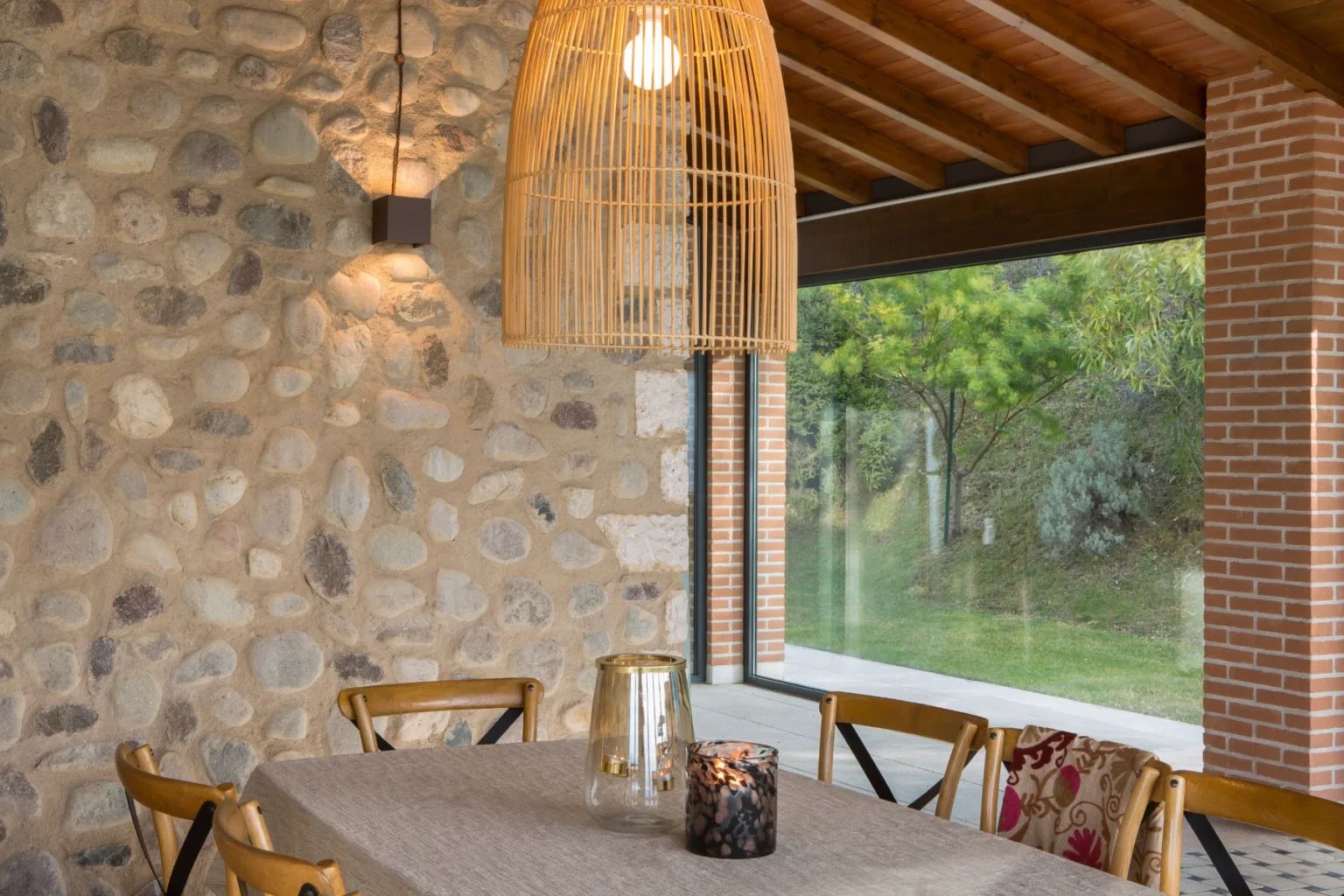Modern dining area featuring a stone wall, wooden ceiling beams, and a stylish woven pendant light, creating a warm and inviting atmosphere. Natural light floods in through large windows, enhancing the contemporary design. | Only on SopranoVillas