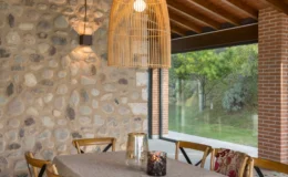 Modern dining area featuring a stone wall, wooden ceiling beams, and a stylish woven pendant light, creating a warm and inviting atmosphere. Natural light floods in through large windows, enhancing the contemporary design.
