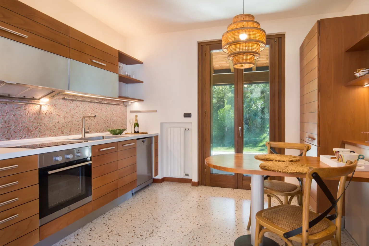 Modern kitchen interior featuring wooden cabinetry, a sleek countertop with a sink, and a stylish pendant light. The space includes a dining table with chairs, a window overlooking greenery, and a mosaic backsplash, creating a warm and inviting atmosphere. | Only on SopranoVillas