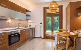 Modern kitchen interior featuring wooden cabinetry, a sleek countertop with a sink, and a stylish pendant light. The space includes a dining table with chairs, a window overlooking greenery, and a mosaic backsplash, creating a warm and inviting atmosphere.