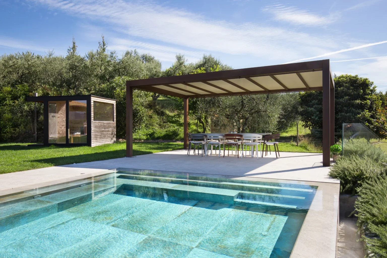 Modern outdoor space featuring a swimming pool, shaded dining area under a pergola, and a wooden garden shed surrounded by lush greenery. Ideal for relaxation and entertaining in a serene environment. | Only on SopranoVillas