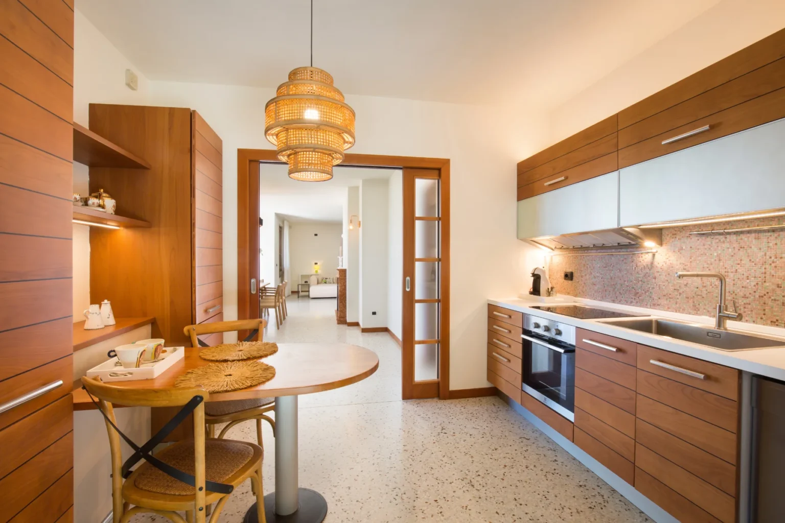 Modern kitchen interior featuring wooden cabinetry, a stylish pendant light, and a cozy dining area with a round table and chairs. The space is bright and open, showcasing contemporary design elements and a clean, functional layout. | Only on SopranoVillas