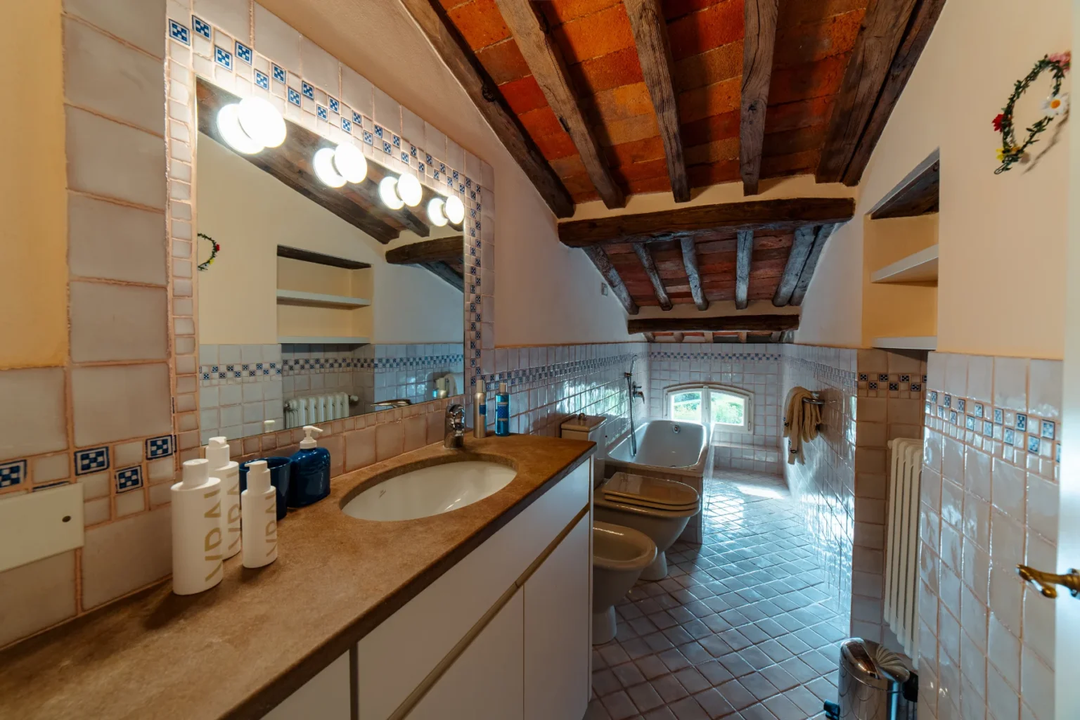 Bright and spacious bathroom featuring a modern sink, bathtub, and bidet, with rustic wooden beams and tiled walls. The well-lit mirror area showcases stylish toiletries, adding a touch of elegance to the design. | Only on SopranoVillas
