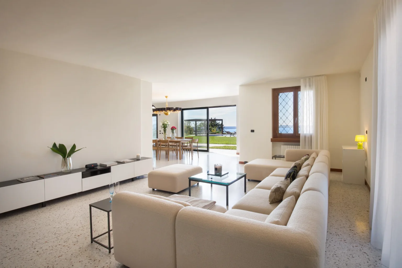 Modern living room with a cream sectional sofa, glass coffee table, and decorative pillows, featuring an open dining area with a view of the outdoors. Large windows allow natural light, showcasing a manicured garden and distant sea view. Elegant decor includes a minimalist sideboard and stylish lighting. | Only on SopranoVillas