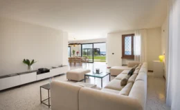 Modern living room with a cream sectional sofa, glass coffee table, and decorative pillows, featuring an open dining area with a view of the outdoors. Large windows allow natural light, showcasing a manicured garden and distant sea view. Elegant decor includes a minimalist sideboard and stylish lighting.
