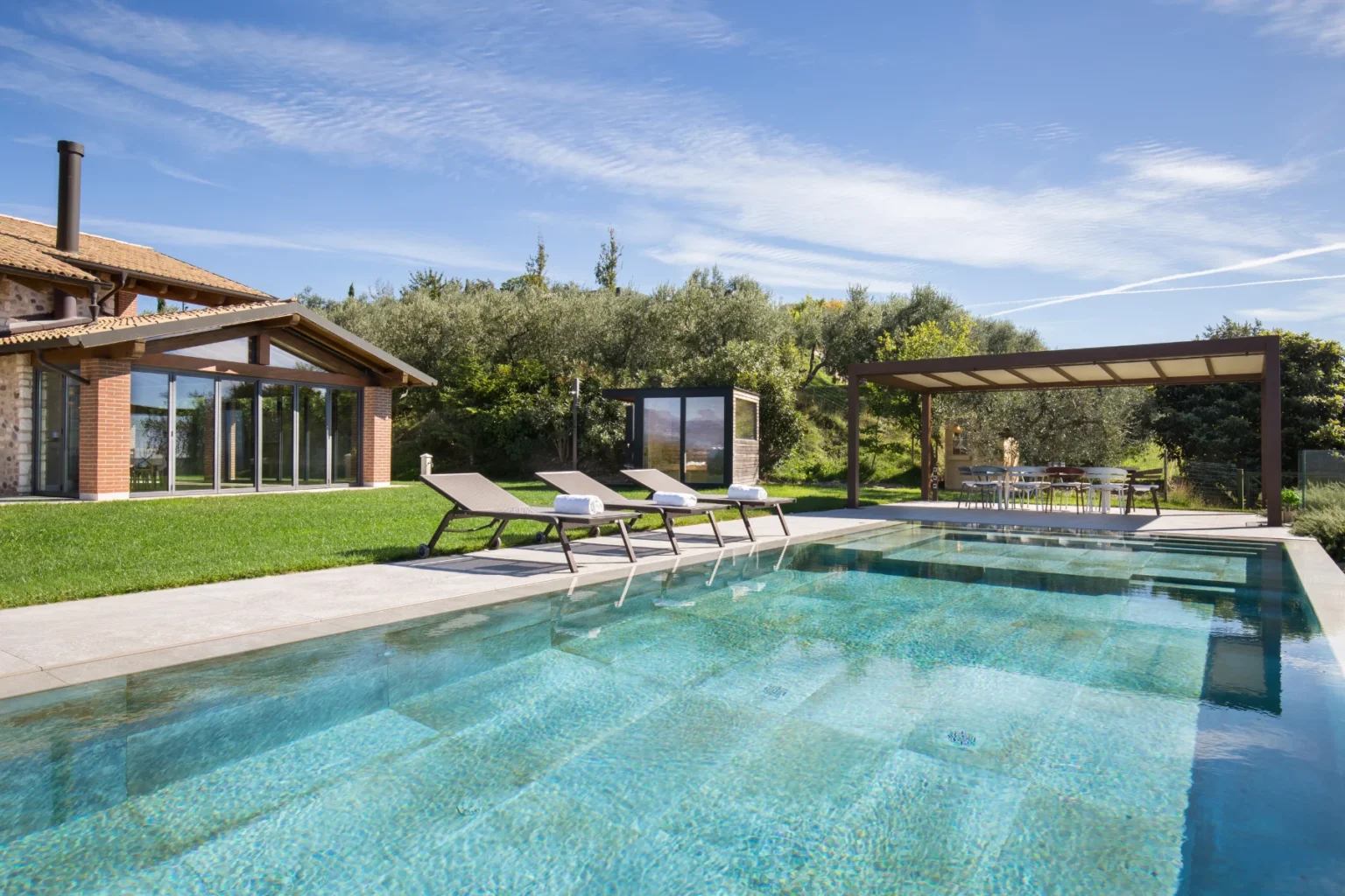 Modern villa with a swimming pool, featuring sun loungers and a shaded dining area, surrounded by lush greenery and a clear blue sky. Ideal for outdoor relaxation and entertaining. | Only on SopranoVillas