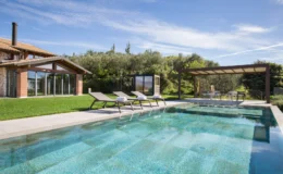 Modern villa with a swimming pool, featuring sun loungers and a shaded dining area, surrounded by lush greenery and a clear blue sky. Ideal for outdoor relaxation and entertaining.