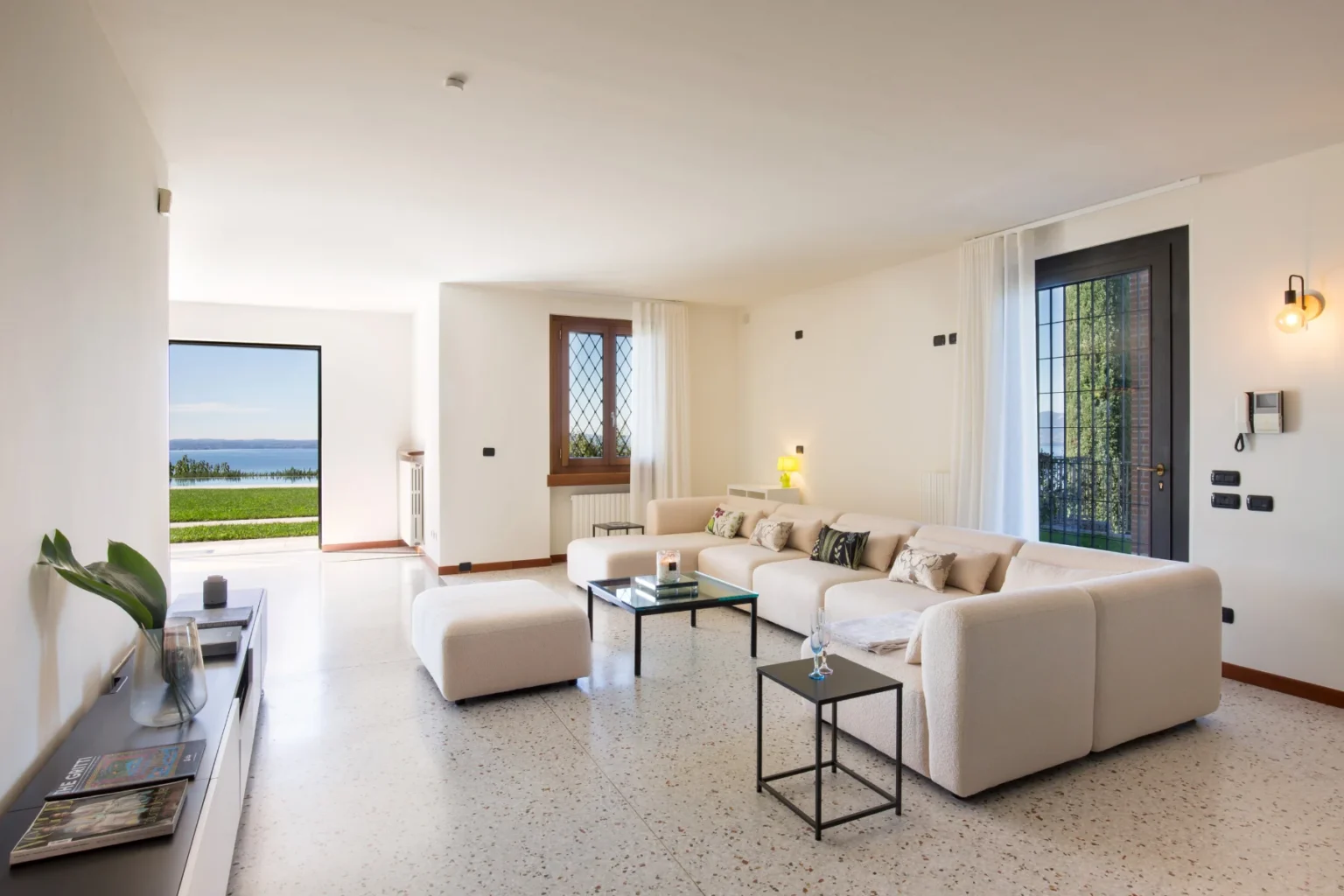 Modern living room featuring a spacious beige sofa, glass coffee table, and large windows overlooking a scenic view. The decor includes a plant and stylish lighting, creating a bright and inviting atmosphere. | Only on SopranoVillas