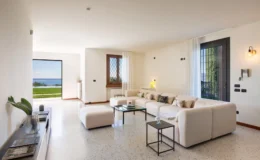 Modern living room featuring a spacious beige sofa, glass coffee table, and large windows overlooking a scenic view. The decor includes a plant and stylish lighting, creating a bright and inviting atmosphere.