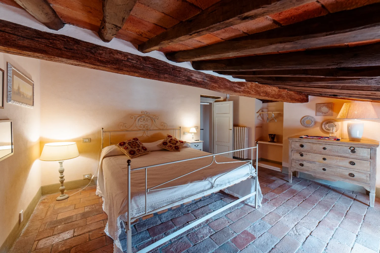 Cozy bedroom featuring a wrought iron bed adorned with decorative pillows, rustic wooden beams, and warm lighting. The room includes a wooden dresser, soft terracotta flooring, and a peaceful ambiance, ideal for relaxation. | Only on SopranoVillas