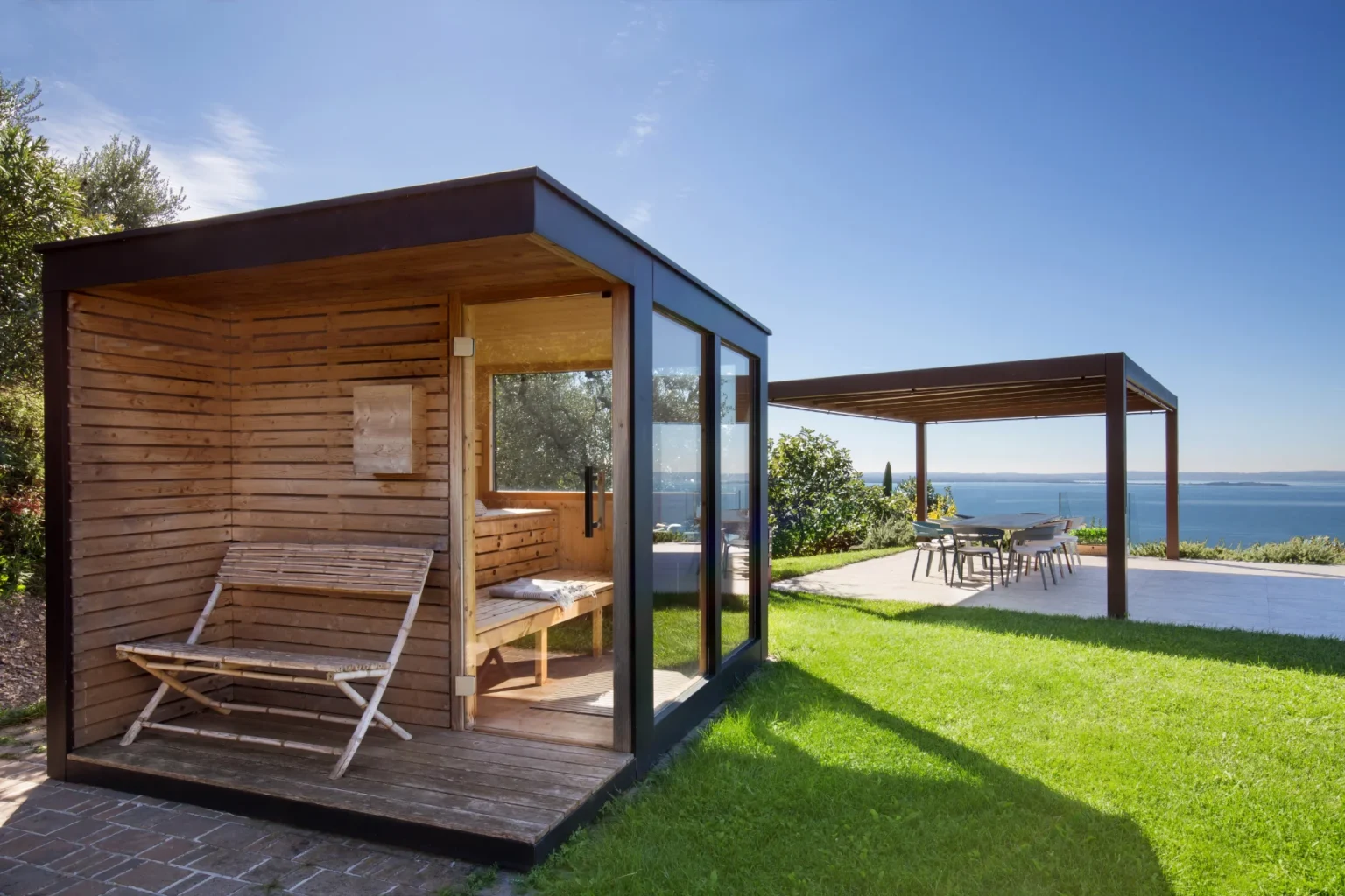 Modern wooden sauna with glass walls and a bench, surrounded by lush green grass and a scenic view of the ocean, complemented by a shaded outdoor dining area. Ideal for relaxation and outdoor living. | Only on SopranoVillas