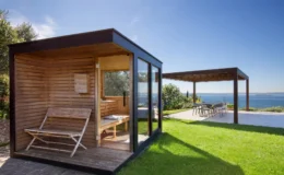 Modern wooden sauna with glass walls and a bench, surrounded by lush green grass and a scenic view of the ocean, complemented by a shaded outdoor dining area. Ideal for relaxation and outdoor living.