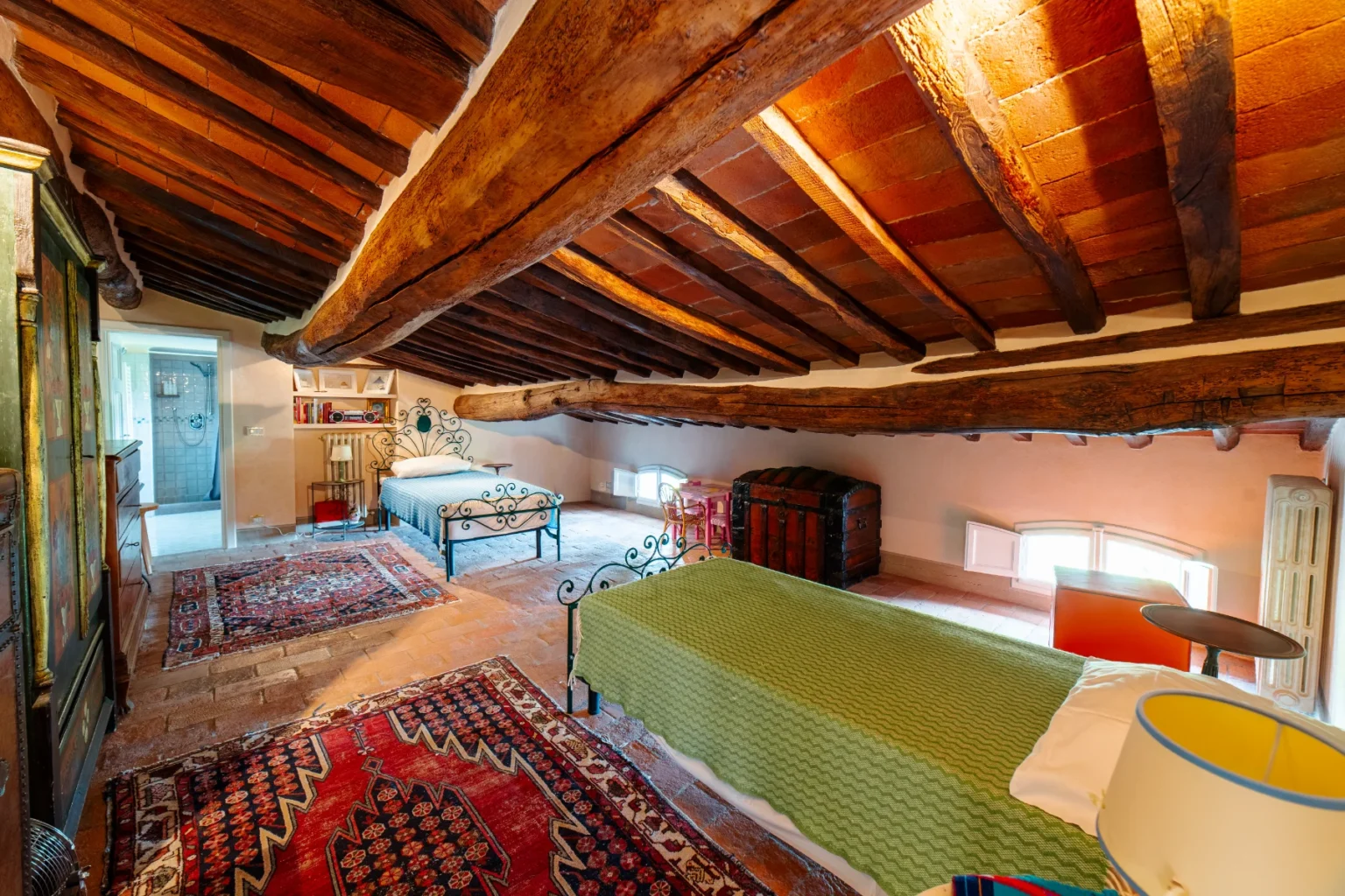 Cozy attic bedroom featuring two iron-framed beds with colorful quilts, exposed wooden beams, and traditional rugs. The room includes a vintage trunk, a small table, and a glimpse of a modern bathroom. Ideal for a charming and rustic getaway. | Only on SopranoVillas