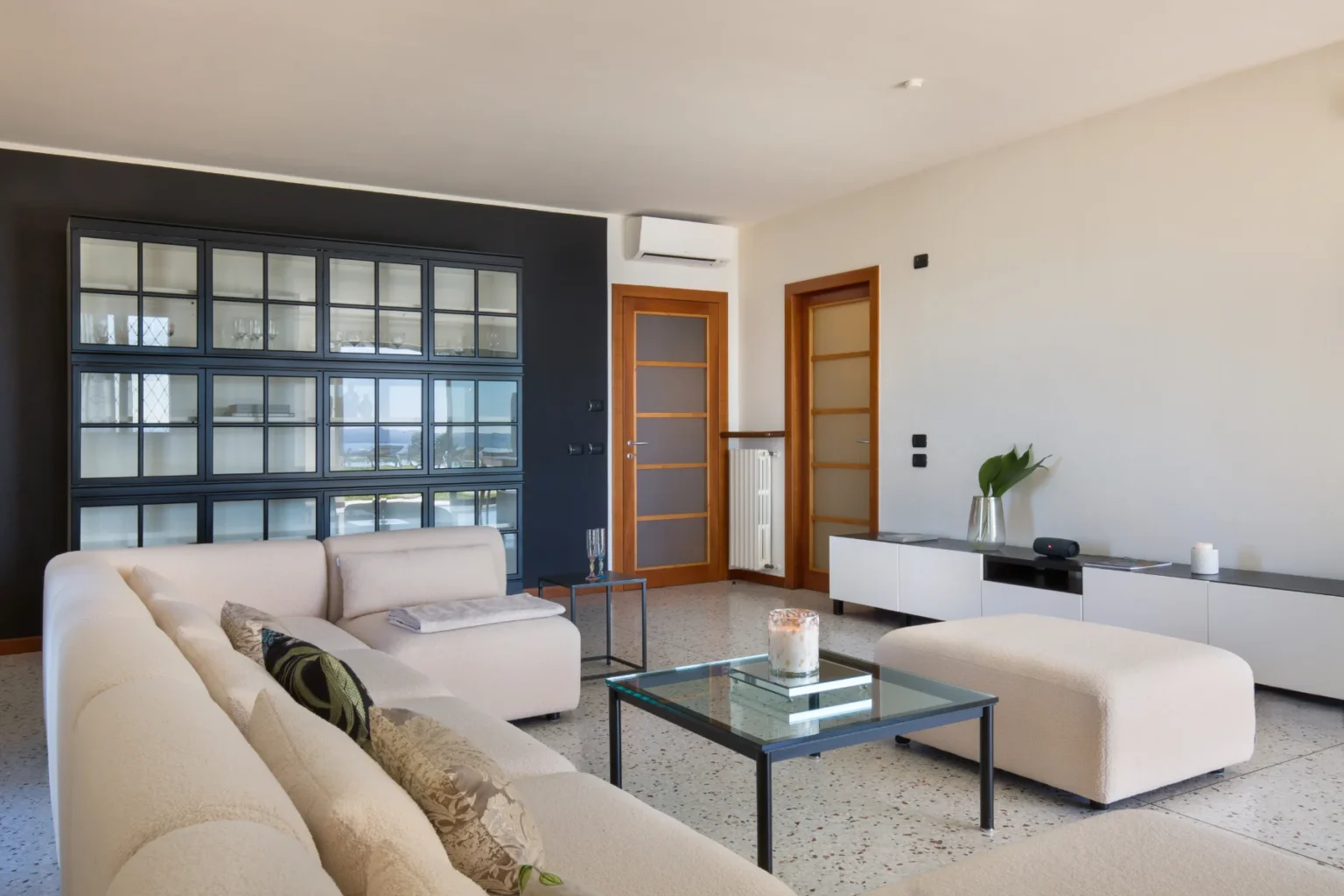 Modern living room featuring a light-colored sectional sofa, minimalist glass coffee table, and a stylish black and glass display cabinet. The space is complemented by wooden doors, a sleek white media console, and decorative plants, creating a contemporary and inviting atmosphere. | Only on SopranoVillas