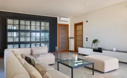 Modern living room featuring a light-colored sectional sofa, minimalist glass coffee table, and a stylish black and glass display cabinet. The space is complemented by wooden doors, a sleek white media console, and decorative plants, creating a contemporary and inviting atmosphere.
