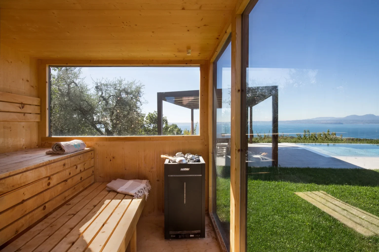 Interior view of a wooden sauna with benches, featuring a black heater and large windows showcasing a scenic view of the ocean and outdoor pool area. The space is designed for relaxation, surrounded by greenery and natural light. | Only on SopranoVillas