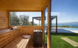 Interior view of a wooden sauna with benches, featuring a black heater and large windows showcasing a scenic view of the ocean and outdoor pool area. The space is designed for relaxation, surrounded by greenery and natural light.