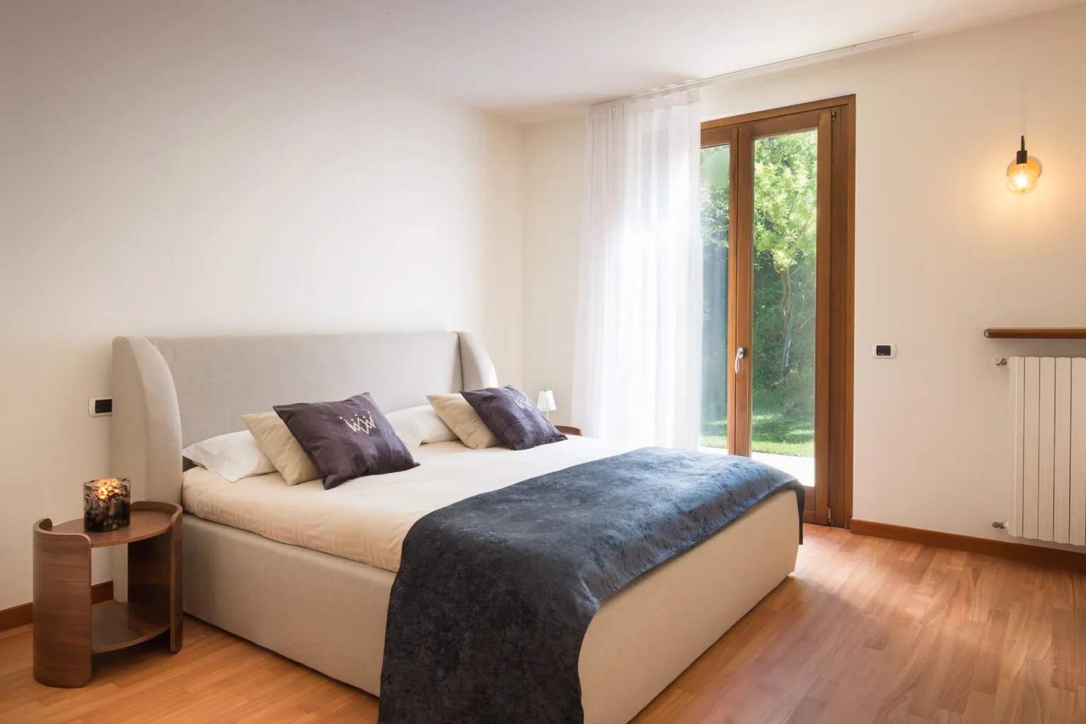 Modern bedroom featuring a comfortable bed with decorative pillows, a cozy throw blanket, a stylish wooden nightstand with a candle, and large windows that provide natural light and a view of greenery. | Only on SopranoVillas