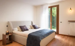 Modern bedroom featuring a comfortable bed with decorative pillows, a cozy throw blanket, a stylish wooden nightstand with a candle, and large windows that provide natural light and a view of greenery.