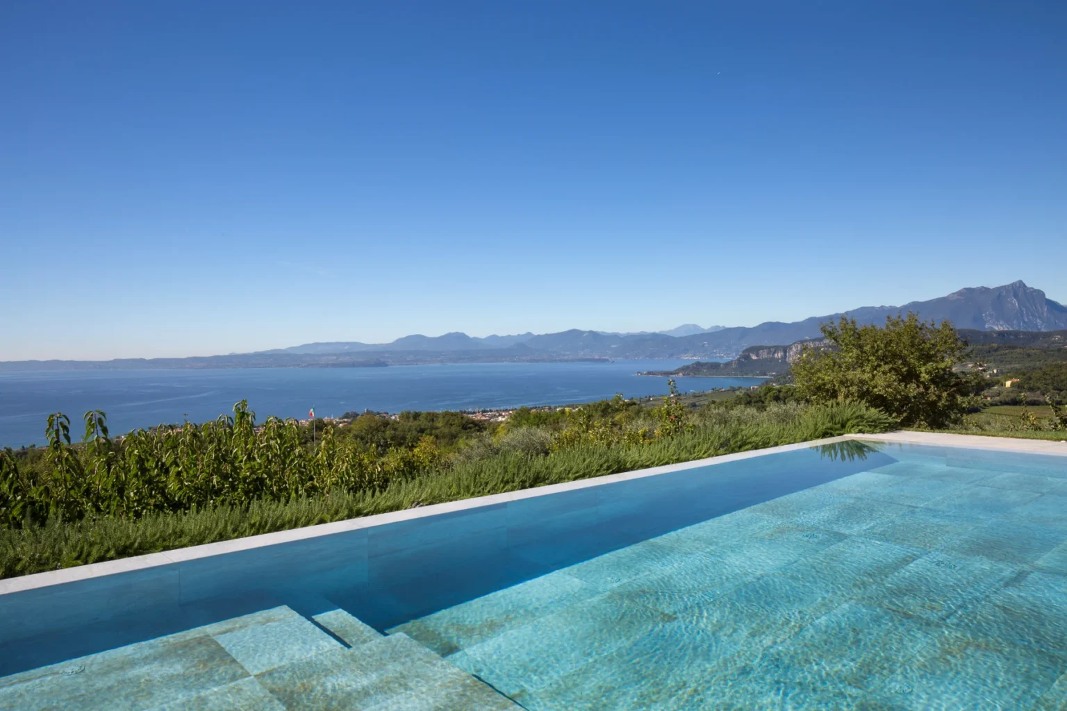 Stunning view of a modern infinity pool overlooking a serene lake and mountainous landscape under a clear blue sky, surrounded by lush greenery. Ideal for luxury vacation retreats and scenic getaways. | Only on SopranoVillas